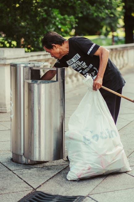 A middle-aged man with short dark hair, dressed in a black T-shirt, is seen leaning forward and placing a white large rubbish bag into a silver, cylindrical public litter bin on a paved walkway. The b