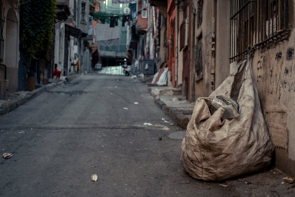 A middle-aged man with dark hair, dressed in a black t-shirt with white text and black trousers, is seen bending slightly while disposing of a white plastic bag filled with rubbish into a tall, cylind
