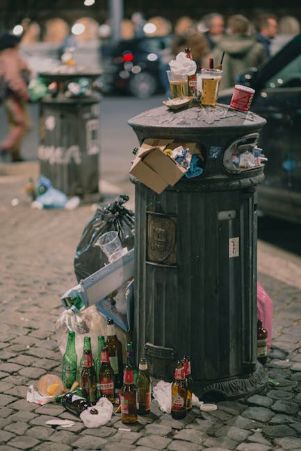 A large pile of mixed waste and rubbish bags, including black plastic garbage bags, flattened cardboard boxes, and various paper and packaging materials, is overflowing from a central grey wheelie bin